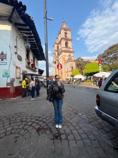 A street scene features a tall church tower with festive decorations in the background. A woman in a leather jacket and jeans stands on a cobblestone street. Buildings line the left side, including a restaurant with signage. People are gathered in the area beyond the woman, under umbrellas and trees. Part of a silver vehicle is visible in the upper right corner.
