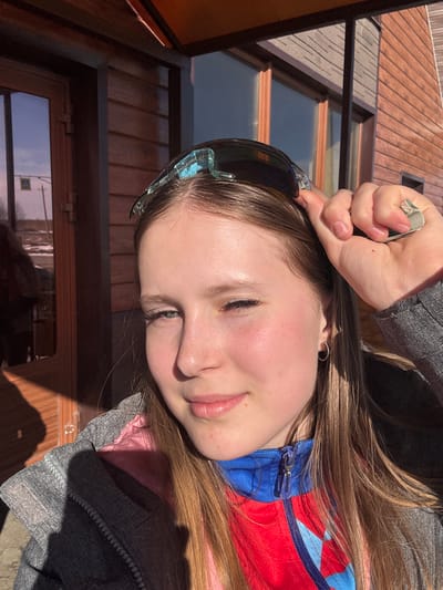 A close-up portrait of a young woman with long brown hair is taken outdoors in Gory, Russia. She is wearing ski goggles pushed up on her forehead and a multi-layered jacket consisting of pink, red, and blue sections, topped with a gray outer layer. Her hand is resting on her forehead, holding a small, square object. The background features wooden walls with windows and the edge of a roof. The image is rotated sideways.