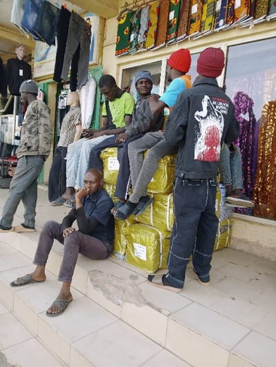 The image depicts an outdoor commercial area in Mumbwa, Zambia. Six males and multiple mannequins are present in front of a storefront. In the immediate foreground, one male sits on a light-colored tiled step, dressed in a dark blue short-sleeved shirt over a black long-sleeved shirt, dark brown trousers, and olive-green sandals, with his right hand resting near his chin. To his right, a stack of approximately twelve large, rectangular packages wrapped in shiny yellow material is arranged, serving as seating. Three males are seated on these packages: from left to right, one male in a light green short-sleeved shirt and light blue jeans; one male in a grey hooded sweatshirt and grey trousers, wearing black sandals; and one male in a light blue short-sleeved shirt and light grey trousers, wearing a red beanie, facing to the right. A fourth male stands with his back to the viewer, positioned to the right of the packages, wearing a black denim jacket with a white and red graphic on the back (partially visible text includes "WE LIVE & WORK... WITHOUT"), dark blue jeans, and dark sandals with socks, also wearing a red beanie. To the far left, a male in a camouflage-patterned hoodie and grey trousers is visible near multiple mannequins displaying dark trousers and shirts. The storefront behind them features a light-colored facade with windows that display colorful patterned fabrics. Various clothing items, including jeans, are hanging from a bar above the mannequins. The ground consists of light-colored tiles, with a damaged concrete step edge visible in the foreground.