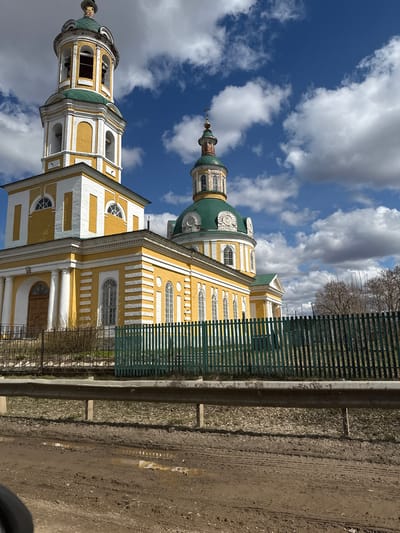 The media file captures a large, ornate Eastern Orthodox church, primarily yellow with distinctive white architectural trim, set against a bright, partly cloudy blue sky. The church features multiple green domes, including a prominent bell tower on the left and a central domed structure topped with a spire and cross. White pilasters, cornices, and decorative frames adorn the walls around numerous tall, arched, and semicircular windows.
The scene is illuminated by clear daylight, suggesting mid-day or afternoon. Bare trees are visible in the background, hinting at an early spring or late autumn season. In the foreground, a dark green slatted fence runs along the right side, separating the church grounds from an unpaved, muddy dirt road. A concrete barrier or guardrail lies between the fence and the road, which shows tire tracks and patches of mud. A small, dark number "3" is visible above an arched doorway on the lower part of the church. No people or specific activities are depicted in the static shot. The architecture and rural-like setting are consistent with the location context of Чутырь, Russia.