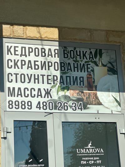 The image shows the facade of a building with a sign for a spa and fitness center. The sign, in large Cyrillic letters, advertises "CEDAR BARREL," "SCRUB," "STONE THERAPY," and "MASSAGE." Below this text is a phone number: "8989 480 26 34." The sign also features an image of spa elements, including candles, stones, and a white towel.
Below the main sign, partially visible through a glass door, is a logo and text for "UMAROVA," a fitness and children's gymnastics studio. This text indicates operating days: "MON - WED - FRI." The building appears to be in Miatli, Russia, as indicated by the location context. The overall scene is a commercial storefront with signage for health and wellness services.