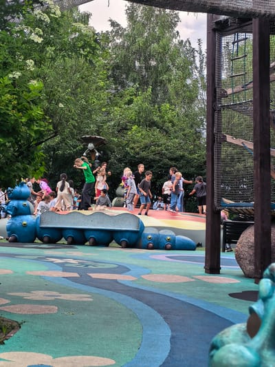 A group of children are playing on a vibrant, multi-colored playground with a large, blue caterpillar-shaped structure. The playground features soft, patterned flooring in shades of green, blue, and beige, with flower-like shapes scattered across it. The blue caterpillar is a prominent play element, with multiple segments and large, round "eyes."
In the background, lush green trees form a dense canopy, suggesting an outdoor, park-like setting. A tall, wooden and mesh climbing structure is visible on the right side of the image, partially obscured by trees and the foreground playground equipment. The sky is overcast, indicating a cloudy day.
The children are engaged in various activities: some are standing on the caterpillar, others are walking or running. One child in a green shirt is in mid-air, appearing to jump or leap. A cartoonish, bug-like figure with an umbrella is mounted on a pole behind the caterpillar. The overall scene depicts a lively and playful environment designed for children's recreation. The specific location of Sovkhoza imeni Lenina, Russia is not visually determinable from the image itself.