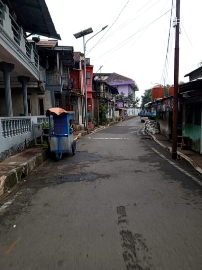 This image depicts a street scene in Pelwehera, Sri Lanka, characterized by a narrow, wet asphalt road lined with buildings. On the left side of the road, a grey concrete building with ornate railings and pillars is visible. Adjacent to it, a series of multi-story structures with balconies and corrugated metal roofs are seen, displaying a mix of faded paint and weathered materials. A blue food cart, possibly selling fried chicken, tofu, and coffee, is parked on the sidewalk in the foreground. The cart has an orange canopy and appears to be made of metal and wood. On the right side of the road, a more modest collection of buildings includes structures with green and rust-colored facades, corrugated metal roofs, and visible electrical wiring. Two large orange water tanks are perched atop one of these buildings. A wooden stool is placed outside one of the establishments. A utility pole with numerous wires runs along the right side of the street. Further down the road, a dark-colored truck is parked, and piles of what appear to be stored materials are visible on the sidewalk. The sky is overcast, suggesting a cloudy day. The overall impression is one of a densely populated urban area with a utilitarian and somewhat aged infrastructure.