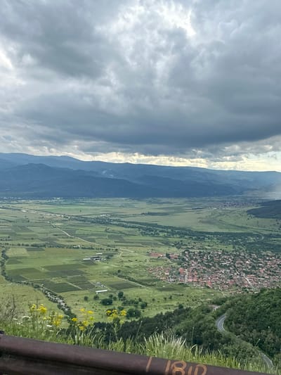 This panoramic landscape photograph captures a vast valley flanked by mountains, viewed from an elevated perspective. The sky occupies the upper portion, featuring a mix of bright, sunlit clouds and darker, heavier cloud formations, suggesting dynamic weather.
A prominent mountain range stretches across the mid-ground, appearing hazy and blue. Below these mountains, the valley floor unfolds into a patchwork of verdant green agricultural fields and pastures. Several settlements are scattered throughout the valley; a noticeable village with numerous reddish-brown rooftops is nestled on the right side, bordered by forested hills. A winding, paved road can be seen traversing the wooded terrain towards this village. Further into the valley, distinct rectangular farm plots and winding paths or small rivers are visible, along with some clusters of white structures, possibly farm buildings.
In the immediate foreground, on the right side of the image, a dark, metallic barrier or railing is present. Tall blades of grass and clusters of vibrant yellow wildflowers grow around it. Faint yellow markings are visible on the barrier, clearly showing the numbers "18" followed by what appears to be a "0" or "D" (e.g., "180" or "18D").
The image depicts a serene, rural setting during daytime, with varied lighting indicating a mix of sun and clouds. The scene conveys a sense of natural beauty and human presence within a mountainous environment, specific to Karnare, Bulgaria.