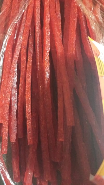 The image shows a close-up view of a bag of dried red meat sticks, likely jerky. The sticks are long and thin, with a slightly rough texture. They are packed tightly together, obscuring much of the bag's packaging, though a sliver of yellow and white label is visible on the right side. The lighting is somewhat dim, creating shadows and highlighting the glossy sheen on some of the meat sticks. The overall impression is of a food product ready for consumption. The location in the city of Юдино, Russia, is not discernible from the image itself.