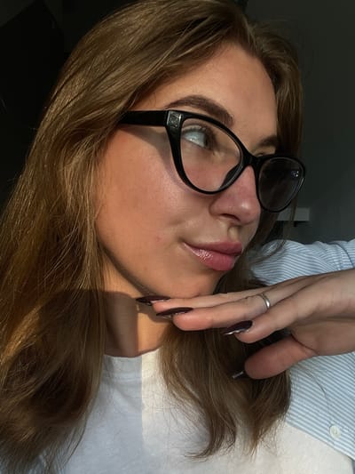 A young woman is captured in a close-up, side profile shot, her head tilted back. She is wearing large, black-rimmed glasses that partially obscure her light-colored eyes. Her lips are glossed and slightly parted. Her hair, a medium brown shade, is styled loosely around her face and shoulders. One hand, with long, dark nails, is raised to her face, fingers delicately touching her cheek, and a silver ring is visible on her index finger. She is wearing a light blue and white striped shirt. The lighting suggests it is daytime, with a strong light source casting shadows across her face, indicating sunlight. The overall mood is contemplative or perhaps a moment of rest. No visible text is present in the image. The setting appears to be indoors, possibly near a window due to the strong directional light and shadows. The location context is Bolesławiec, Poland.