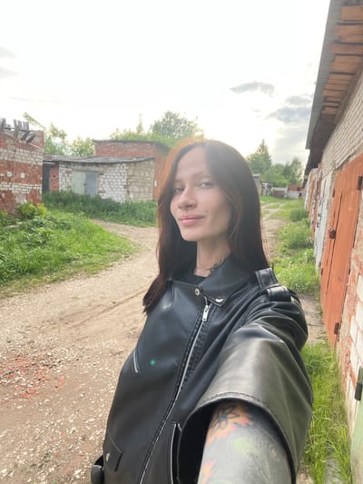 A person with long brown hair and a black leather jacket is standing on a dirt path. The person has a pierced nose, visible tattoos on their neck and arm. The background features brick buildings with closed garage doors and overgrown vegetation. The location is inferred from the metadata to be in Oktyabr'skii, Russia.