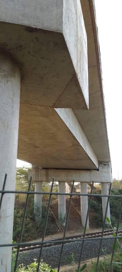 A low-angle shot reveals a concrete bridge supported by cylindrical pillars. A section of the bridge juts out, creating a triangular overhang. Below the bridge, smaller pillars are visible, supporting a lower tier. A railway track runs beneath the bridge. A wire fence with foliage obscures part of the foreground. Patches of green vegetation and trees are also visible.
