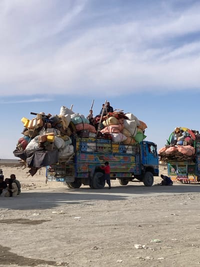 The image depicts two heavily overloaded, brightly decorated trucks on a dirt and gravel expanse under a partly cloudy blue sky in Chaman City Tehsil, Pakistan. The primary subject is a blue-cab flatbed truck, adorned with multicolored, folk-art style paintings, stopped or parked. Its cargo bed is piled high with a precarious, voluminous load consisting of numerous large sacks (various colors including white, pink, green), yellow and other colored plastic containers, bundles, wooden planks, and assorted household items, secured with ropes and tarpaulins.
Approximately five individuals, including at least two adults and three children, are visible seated or standing atop this cargo load. A young boy in a red shirt and dark pants stands on the ground near the front passenger wheel of this truck. To the far left in the foreground, two adult males in traditional attire are seated on the ground. Further back and to the right, a second, similarly overloaded and decorated truck is partially visible, with one individual crouching on the ground beside it. The environment is arid, characterized by dry, light-colored soil and an expansive, clear horizon with a faint distant structure. The sky is light blue with scattered white, wispy clouds.