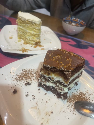Two slices of cake are presented on white plates. The slice in the foreground is a dark chocolate cake with layers of cream and a chocolate ganache topping sprinkled with nuts. It is placed on a white plate dusted with cinnamon. The second slice, further in the background, appears to be a lighter colored cake, possibly vanilla or sponge cake, with multiple layers and white frosting. A small decorative bowl with a blue and orange pattern is visible on the right side of the image, situated on a wooden surface. The table is adorned with a tablecloth featuring a bold, abstract design in pink and dark blue. The scene suggests a casual dining setting, possibly in a cafe or restaurant in Atyrau, Kazakhstan.