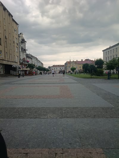 The image shows a view of a paved pedestrian area in front of buildings in Drohobych, Ukraine. The paving is made of large, grey flagstones with rectangular sections of pinkish-orange and smaller grey cobblestones interspersed. The area is lined with trees and a grassy area with benches. Several people are walking in the distance on the pedestrian area and some are seated in an outdoor cafe. The buildings on the left are multi-story with a light yellow facade and windows. The buildings across the street are of various architectural styles and colors, with pitched roofs. The sky is overcast with grey clouds.