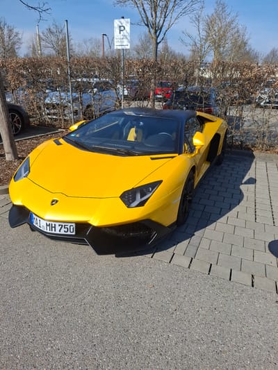 This image captures a bright yellow Lamborghini Aventador, identified by its German license plate "IN AI MH 750," parked on a sunny day in Ingolstadt, Germany. The vehicle is situated with its front tires on asphalt and its right side and rear tires on rectangular grey pavers. It features a black convertible top and a black interior with yellow accents. In the background, a leafless hedge separates the immediate parking area from a larger lot containing numerous vehicles, primarily Audi models. A white sign displaying the Audi rings logo and the text "P Parken zum Neuwagen abholen ->" (Parking for new car pickup) is visible behind the hedge, indicating the location is likely at the Audi Forum Ingolstadt or a related Audi facility. Bare trees and multiple industrial chimneys or stacks are present on the distant horizon. Direct sunlight illuminates the scene, casting prominent shadows to the right and rear of the Lamborghini. No people or specific interactions are visible within the frame.