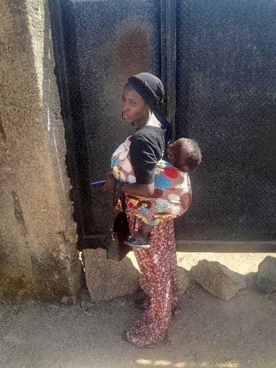 The image captures an adult female carrying an infant on her back, standing outdoors in Barakin Acha, Nigeria. The female, positioned center-frame, wears a black headwrap, a black short-sleeved top over a patterned undergarment, and a long, reddish-brown floral-patterned skirt. She holds a dark blue object, consistent with a mobile phone, in her left hand and a dark handbag hangs from her left shoulder. The infant, secured to her back with a multicolored fabric wrap featuring circular patterns, wears dark shoes and is partially visible with its head turned to the left. To the immediate left is a light-colored, roughly textured stone pillar or wall section. The background consists of a dark, weathered metallic surface, possibly a corrugated gate or wall, exhibiting reddish-brown discoloration consistent with rust. The ground is dry, light-brown soil, with several light-colored rocks and boulders scattered at the base of the structures. The scene is illuminated by direct sunlight from the right, casting shadows.