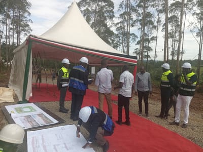 This outdoor photograph captures a group of men gathered at what appears to be a project site in Moticho ward, Kenya. A large, white pyramidal tent, festooned with fabric stripes resembling the Kenyan flag (green, red, black, and white), dominates the left side of the frame. A red carpet extends from beneath the tent, leading to where the individuals are assembled.
Around ten to fifteen men are visible, most of whom are wearing white hard hats and high-visibility safety vests, either yellow or blue with black details. One man in a white hard hat and blue vest is crouched in the foreground, examining large architectural blueprints and plans laid out on the red carpet. Other men stand nearby, some also in safety gear, and others in casual attire like striped or solid-colored shirts, seemingly engaged in discussion or reviewing documents.
The background is characterized by numerous tall, slender trees, likely eucalyptus, stretching towards a cloudy or overcast sky, indicative of a natural or semi-rural setting. The activity suggests a site visit, project briefing, or possibly a groundbreaking ceremony, with participants focused on the development plans.
Visible text includes "OLDINGS LTD" on a blue safety vest. The blueprints display various architectural drawings, floor plans, and some smaller, illegible textual labels. One plan clearly illustrates green areas, possibly indicating landscaping or recreational spaces. The scene takes place during daytime, under a bright, diffused light.