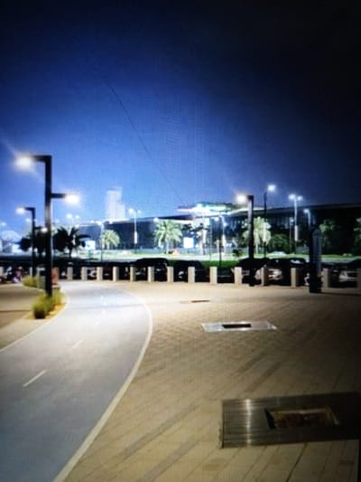 This media file depicts an outdoor scene at night, likely in Golbembal, Nigeria. The primary subjects are a pathway and a building complex in the background. The pathway is paved with light-colored tiles and features a darker, possibly asphalt, lane for cycling or walking, marked with white lines. Streetlights, with their warm glow, illuminate the pathway and the foreground area.
In the mid-ground, a row of pillars suggests a covered walkway or an entrance to the building. Beyond these pillars, a large, modern building is visible, characterized by its dark facade and illuminated windows. Palm trees are scattered in front of the building, indicating a potentially tropical or subtropical climate. There are some vehicles parked near the building, suggesting a public space like an airport, a convention center, or a large shopping complex.
The activity appears to be ambient, with no clear indication of a specific event. The presence of people in the far left of the image, blurred and indistinct, suggests some level of public access and activity, though the overall mood is quiet and serene. The time of day is clearly night, with a deep blue sky and artificial lighting dominating the scene. The weather appears clear.
Notable details include the modern architectural style of the building, the well-maintained pathway, and the lighting scheme that creates a welcoming atmosphere. There is no visible text in the media file. The image conveys a sense of order and modern development.