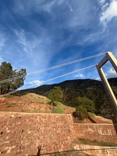 A low-angle photograph, taken outdoors, presents a landscape featuring a red rock mountain face and a clear blue sky. Dominating the top of the frame is a section of a bridge or similar structure made of light-colored concrete. Thin, white cables run vertically from the structure down toward the bottom of the image.
To the right of the frame, a wall constructed of reddish-brown stone is visible. Vegetation, including trees and bushes, grows along the cliff face of the mountain, adding a touch of green to the scene. The sky above is a vibrant blue dotted with scattered white clouds.
This image is situated in Tabaroucht, Morocco.