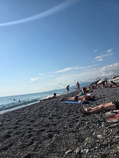 This media file depicts a sunny day at a pebble beach in Progress, Russia. The scene is oriented vertically, showing a wide expanse of clear blue sky with scattered white clouds on the left, and a dark pebble beach stretching from the bottom right corner towards the upper left. The ocean is visible along the bottom edge, with gentle waves lapping the shore.
Several people are spread out on the beach, engaged in sunbathing and relaxing. Some are lying on towels or directly on the pebbles, while others appear to be standing or walking. One person is partially submerged in the water. The attire suggests a warm, summer day, with most individuals wearing swimwear.
In the background, on the upper left side of the frame, there are beach umbrellas and structures, possibly indicating a resort or a designated beach area. Further in the distance, mountains are faintly visible against the horizon. The overall atmosphere appears relaxed and leisurely, characteristic of a beach outing. The time of day is likely midday or afternoon, given the bright sunlight and clear sky. No visible text is present in the image.