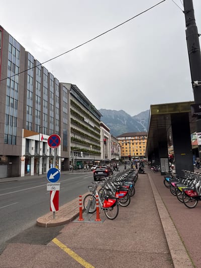 The image depicts a street scene in Innsbruck, Austria, with a prominent view of the Alps in the background. The foreground features a row of rental bicycles lined up along a sidewalk. These bicycles have red and white branding, with some displaying web addresses like "lvb-jobs.at".

To the left of the bicycles is a sidewalk with traffic signs. One sign is circular with a red border and a diagonal red line, indicating no parking, and reads "ANFANG". Below it, a blue circular sign shows a white arrow pointing left, and a rectangular sign below that reads "ausgenommen" and displays a bicycle symbol.

Behind the sidewalk, a street is visible with cars, pedestrians, and a traffic light. On the left side of the street, a modern building with a grey facade and numerous windows stands tall. A bank with the "BAWAG" logo is present on the ground floor of this building.

Further down the street, another building with a yellow facade and many windows is visible. The backdrop is dominated by the majestic Innsbruck Alps, with their peaks partially obscured by a cloudy sky. The overall environment suggests an urban setting on an overcast day.