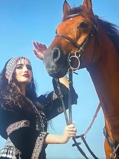 In the city of Shakhovskaya, Russia, a woman with dark wavy hair, adorned with a beaded headdress and wearing a black garment with intricate silver and red embroidery, is standing next to a bay horse. The woman is gently touching the horse's face with her left hand, her fingers adorned with rings. Her right hand holds the horse's bridle. The horse is wearing a brown leather bridle with a silver bit. The background is a clear, bright blue sky. The scene appears to be an outdoor setting.