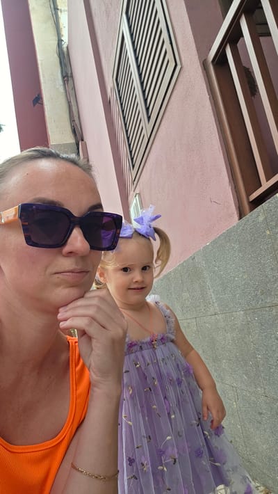 In the city of Guía de Isora, Spain, a medium shot captures a woman and a young child outdoors. The woman, positioned on the left, wears an orange sleeveless top and large purple sunglasses. Her right hand is raised to her chin, and she has a neutral expression. To her right, the young child is dressed in a lilac tiered dress adorned with floral embroidery. The child has blonde hair styled in two pigtails, each secured with a purple bow, and wears a pink pendant necklace. The background features a building with a pink facade, featuring a diamond-shaped window shutter and a wooden railing. A grey stone wall runs along the lower portion of the image. The lighting suggests it is daytime, with diffused natural light.