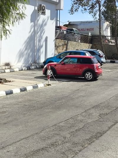 The image captures an outdoor scene in Kızılbaş, Northern Cyprus / Cyprus, under bright daylight conditions. A red two-door Mini Cooper is centrally positioned, parked with its front bumper area connected by a metal chain to a red and white striped bollard. Behind it, a blue sedan and a light blue/grey SUV are parked in sequence. To the left, a white building wall with an externally mounted air conditioning unit shows shadows from unseen foliage. A curb with alternating black and white painted sections separates the parking area from a paved path. In the background, a stone-faced retaining wall supports a metal railing, beyond which a large white tanker truck with "TEHLİKELİ MADDE" (Dangerous Material) and "YAKIN DOĞU AKARYAKIT" (Near East Fuel) visible text is partially obscured by pine trees. The asphalt ground exhibits various textures and tire marks. No individuals are present in the scene.