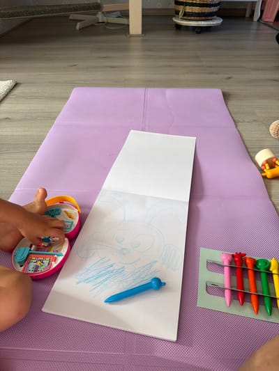 A young child's left leg and hand are visible on the bottom left of the frame, positioned on a purple mat. The child is playing with a pink toy with a yellow handle, which appears to be a miniature dollhouse. Next to the toy is a white sketchbook with a blue crayon drawing of a bunny. A blue crayon rests on the drawing. To the right of the sketchbook, a green holder contains five colorful pens with animal-shaped tops: pink cat, red deer, orange fox, green bear, and yellow lion. A yellow toy with orange beads is visible on the purple mat to the right of the pens. The background shows a wooden floor and furniture, including a woven basket on a wheeled stand. The scene appears to be indoors, likely in a home in Melick, Netherlands.