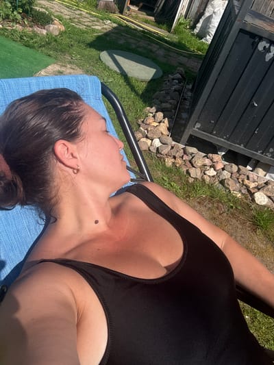 An adult female is lying on a blue lounge chair outdoors, with her head resting on her arm. She is wearing a black one-piece swimsuit. Her eyes are closed, and her face is tilted upwards, suggesting she is basking in the sun. The background shows a grassy area with scattered rocks, a green turf mat, and a dark, corrugated metal structure. The lighting indicates it is daytime, likely sunny. The scene appears to be a residential backyard. There is no visible indication that this location is in the city of Petushki, Russia.