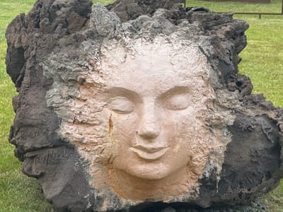 The image shows a large, roughly textured rock formation that has been sculpted to depict a human face. The face, rendered in a lighter, smoother material, is centrally located on the rock, with closed eyes and a gentle smile. The surrounding rock is dark and jagged, with visible crevices and a rough surface. There are some dark, graffiti-like markings on the left side of the rock, below the sculpted face. The rock is situated outdoors, on a patch of green grass, suggesting a park or open space. The background is blurred green grass and a hint of a fence or railing, indicating an outdoor, possibly public setting. The overall impression is that of a natural rock that has been artistically enhanced. The location of Kuril'sk, Russia is not discernable from the image itself.