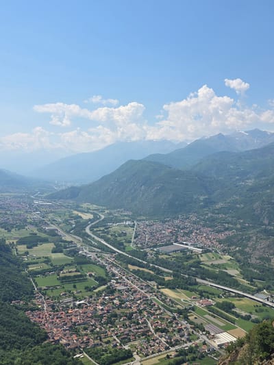 The image shows an aerial view of Sant'Ambrogio di Torino, Italy. Mountains rise on the left of the image, transitioning into a valley filled with a town and green agricultural fields. A highway snakes through the landscape. Clouds fill the top left corner of the image and fade into the clear blue sky. The town is densely packed with buildings featuring red-tiled roofs.