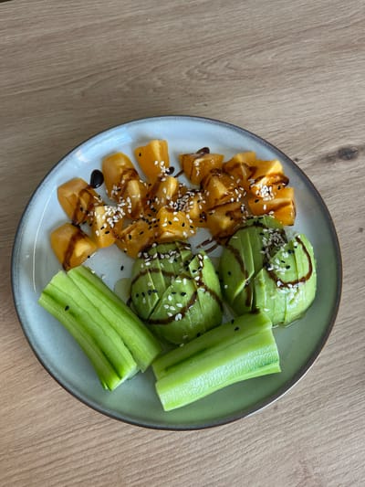The image shows a plate of food. The plate contains sliced persimmon topped with a dark sauce and sesame seeds, sliced avocado topped with the same dark sauce and sesame seeds, and sliced cucumber. The plate is on a wooden surface. The location is unknown, and there is no evidence to suggest it is in Ādaži, Latvia. There are no people visible in the image.