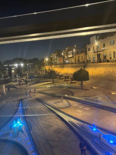 This is a nighttime exterior shot taken from a high vantage point, looking down at a public plaza with a fountain and surrounding buildings. The time of day is clearly night, with dark skies and artificial lighting illuminating the scene. The setting appears to be an urban area, possibly a city square or promenade, given the presence of buildings and paved walkways.

The main subjects are people scattered throughout the plaza, some walking, some standing, and a few gathered around the fountain area. The fountain itself is a prominent feature, with water cascading and illuminated by bright blue lights, creating a striking visual effect. There are also trees and what appear to be art installations or decorative elements within the plaza. The surrounding buildings are multi-story structures, some with windows lit from within, suggesting they are residential or commercial.

One notable detail is a sign visible on one of the buildings that reads "LOZOUDI" in white lettering. The overall mood of the image is serene and perhaps a little magical due to the nighttime lighting and the illuminated fountain. The weather appears to be clear, as there are no indications of rain or strong winds. The composition suggests the photograph was taken from an elevated position, possibly a balcony or window overlooking the plaza.