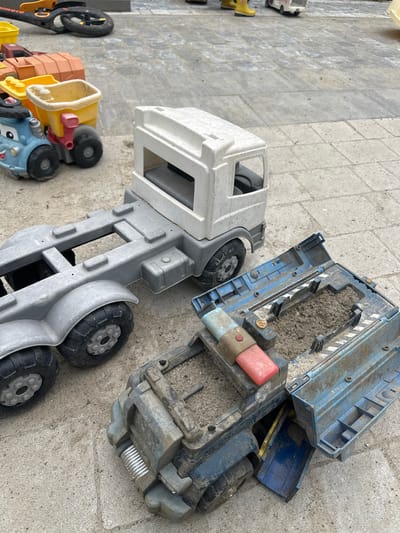 The image shows a collection of dirty, well-used children's toy vehicles on a grey paved surface, possibly a backyard or playground. In the foreground, a blue toy truck with a detached, open cargo bed filled with dirt is partially visible. Next to it is a large, grey toy truck cab and chassis. To the left, a smaller blue toy dump truck with a yellow bed is visible. In the background, other toy elements and a black scooter are scattered. The overall impression is one of outdoor play, with the toys appearing weathered and covered in dirt. The location is stated to be Malinovka, Russia.
