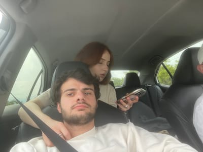 A man and a woman are visible inside of a car. The man is wearing a white sweater and has a seatbelt across his chest. He is holding a phone in his right hand. The woman has shoulder-length auburn hair and is resting behind the man in the seat. The interior of the car is a light grey color. Outside the car is green foliage and glimpses of the sky. The location in the city of Changilari, Georgia cannot be determined.
