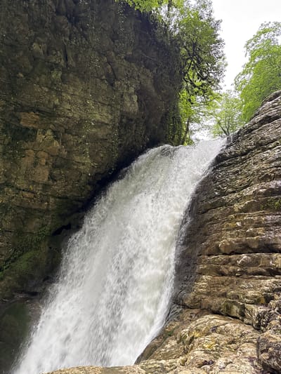 A photograph captures a waterfall cascading down a rugged cliff face. The water appears as a blurred stream, indicating movement. The cliff on the right side of the frame displays a stratified rock formation, with vertical layers and varying textures. Vegetation, including trees and moss, grows on the left cliff face, adding a touch of green to the scene. Above, the sky is visible through the opening between the cliffs, appearing bright and overcast. This image is most likely taken in Tsabal, Abkhazia.