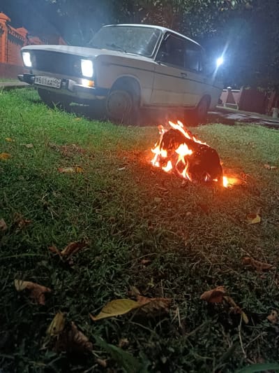 The image depicts an outdoor scene at night, featuring a white sedan car parked on a grassy area. The car's headlights are on. In the foreground, a small fire burns on the grass. Scattered autumn leaves are visible on the ground. In the background, a brick fence and trees can be observed. The scene appears to be in a residential area, possibly a backyard or a park. There are no people visible in the image. The overall atmosphere is dark and somewhat mysterious due to the nighttime setting and the fire. The location is identified as Bratskoye, Russia.