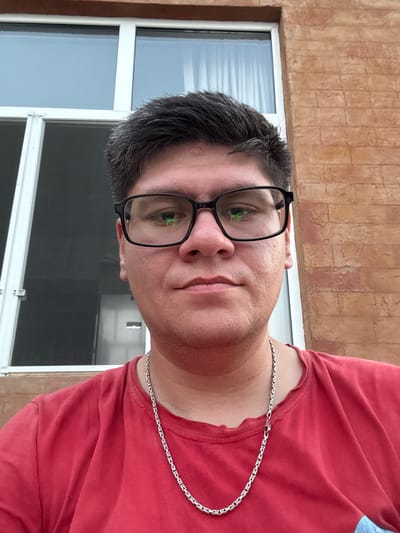 This is a portrait of a person wearing glasses and a red t-shirt, with a silver chain around their neck. The background shows a textured wall and a window with blinds. The lighting suggests it might be daytime. The image is taken from a slightly high angle, looking down at the subject. There is no visible text in the image.