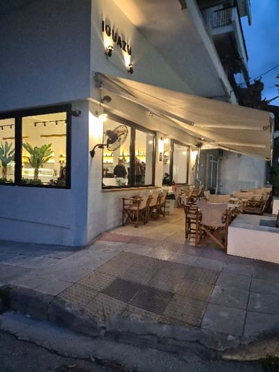 An exterior view depicts the business "IGUAZU" in the Municipal Unit of Corinth, Greece, illuminated in the evening. The establishment features a light-colored building facade with "IGUAZU" prominently displayed and lit by two wall-mounted uplights. A retractable light-colored awning extends over an outdoor seating area on the paved sidewalk. This area contains multiple light-stained wooden chairs with beige fabric seats and backs, arranged around several small tables. A black wall-mounted oscillating fan is affixed to the building's exterior.

Large glass windows offer a view into the brightly lit interior, where several individuals are visible: one person stands behind a counter, and other patrons are seated or standing. Green leafy plants are present inside the left window. The sidewalk consists of grey and reddish-brown tiles, including a tactile paving section with a rectangular metal grate. A concrete curb separates the sidewalk from a darker asphalt street surface. In the background, an upper floor of an adjacent building with a balcony is partially visible against a dark sky.