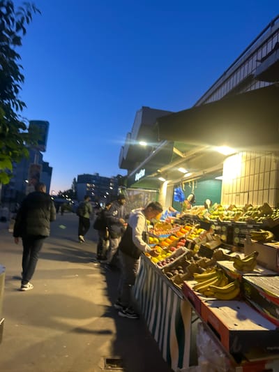The image shows a street scene in Noisy-le-Grand, France, at dusk. A fruit stand is positioned on the right side, stocked with various fruits including bananas, oranges, lemons and avocados. A man is working at the stand, arranging or serving customers. Several pedestrians are walking along the sidewalk in front of the fruit stand. Buildings and trees are visible in the background against the blue sky.