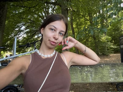A young woman with dark hair and blue eyes is sitting on a weathered wooden bench outdoors, leaning her head on her hand. She is wearing a brown ribbed tank top and a white shell necklace. Her fingernails are painted with bright colors, including coral and turquoise. Behind her, lush green trees and foliage create a dappled light effect. To her left, a bicycle is partially visible. The scene appears to be set in a park or wooded area within the vicinity of Seulberg, Germany.