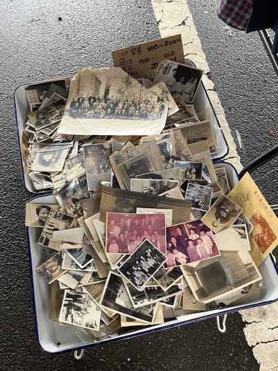 The image shows a close-up, high-angle view of a two-sectioned enamel tray filled with a collection of old photographs. The tray sits on a textured, asphalt-like surface with a painted white line visible, possibly indicating a road or parking area. A portion of a person's plaid-patterned clothing is visible in the upper left corner. The photographs vary in size and orientation. Some are black and white, while others have a sepia or pinkish tone, suggesting aging and possibly different development processes. The photographs depict various subjects, including individual portraits, group photos of people in formal attire, building exteriors, and outdoor scenes. A cardboard tag is visible among the photos, with handwritten Japanese text indicating a price range of 100-500 yen for the photographs and the approximate date of the photos as "1910 ~ OLD". A postcard is also present, with text including "Kanazawa Garden Memories". The scene suggests a vintage photograph sale or display, perhaps at a flea market or antique fair. The location cannot be determined, although it is indicated in the photo contents that the vintage photos are from Kanazawa Garden. The text on the cardboard tag provides clues about the object's purpose, indicating they are for sale.