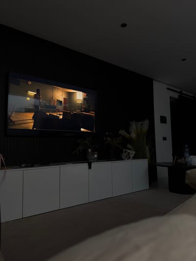 The scene depicts a dimly lit modern entertainment area within a residence in Tassoultante, Morocco. A large flat-screen television is centrally mounted on a dark, vertically textured accent wall. The screen displays a scene featuring a male figure standing in what appears to be an industrial or workshop-like interior, characterized by warm light, a visible wall clock, and various objects including chairs and machinery in the background.

Below the television, a long, white, multi-segment media console extends across the wall. On its surface, several decorative elements are present, including a clear glass vase, potted plants, and what appear to be tall, dried ornamental grasses or pampas grass on the right end. Small items, possibly remote controls, are also visible on the console's left side.

To the right of the media console, a round, dark-ribbed side table holds a plastic bottle of water and other indiscernible objects. The floor is covered with a light-colored carpet or rug. The foreground shows a blurred portion of a light-colored upholstered surface, likely a couch or bed. The room's ceiling features recessed spotlights, and a section of a white wall with a dark sliding barn-door hardware mechanism is visible on the far right. No individuals are directly observed in the room, with the only visible human form being a depiction on the television screen.