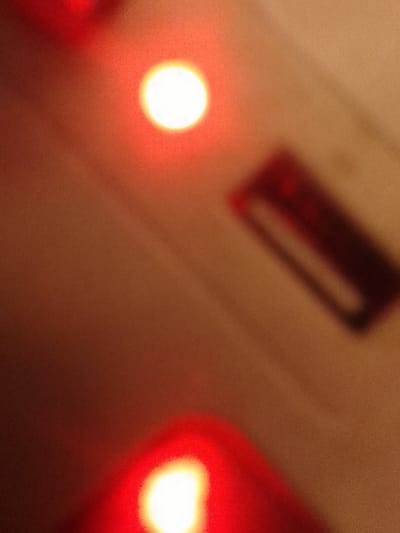 The image depicts a heavily blurred, low-light scene located in Orifite, Nigeria, dominated by warm reddish-brown and red tones. A bright, circular white light source is central in the upper half of the frame, surrounded by a red diffusion. A second, similarly bright white light source, also emanating a red glow, is partially visible at the bottom-center. In the upper-right quadrant, a dark, rectangular object with a lighter internal perimeter is discernible, though its specific features are obscured by blur. No individuals, specific actions, or interactions are identifiable. The background appears as an amorphous field of reddish-brown.