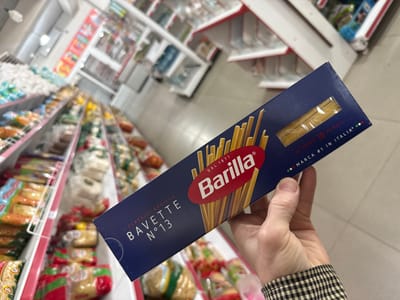 A person holds a box of Barilla Bavette pasta in a grocery store aisle. The box is dark blue with the Barilla logo, which is a red oval with the brand name in white. Text on the box includes "I CLASSICI BARILLA", "BAVETTE N°13", "DAL 1877", "AL DENTE 10 MINUTI", and "MARCA #1 IN ITALIA". The pasta inside the box is visible through a rectangular window. The background shows shelves stocked with various food products, and the lighting suggests an indoor setting, likely a supermarket. The person's hand, wearing a black and white checkered shirt sleeve, is holding the box. The scene is angled, with the camera looking down and to the side.