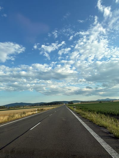 The image depicts a rural road leading into the distance, bordered by agricultural fields. The sky is a vibrant blue with scattered white clouds, some of which are tinged with gray, suggesting a mix of sunlight and cloud cover. The road itself is asphalt with white lane markings. On the left side of the road, there is a field of dry grass or harvested crops, and further back, a line of trees forms a dark border against the horizon. On the right side, a lush green field of crops stretches towards the distance. Gentle, rolling hills are visible in the background, with a few scattered trees and the faint outline of mountains on the far horizon. There are no people or vehicles visible in the image. The overall impression is one of a peaceful, agricultural landscape in daylight. The location is identified as Kožuchov, Slovakia.