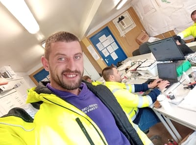 The image captures an indoor office environment located in Drugan, Bulgaria, occupied by four individuals. In the immediate foreground, a light-skinned male with short dark hair and a beard is depicted, smiling directly at the camera. He wears a bright yellow high-visibility jacket over a purple hoodie.

To the right of the foreground subject, a second light-skinned male with short dark hair is seated at a light-colored desk, actively typing on a black laptop. He is dressed in a yellow high-visibility jacket featuring reflective stripes on the arms. A third male is partially visible on the far right, also wearing a yellow high-visibility top. A fourth individual, largely obscured, is seated behind the foreground subject.

The office features multiple light-colored desks, which are cluttered with various items including laptops, an external monitor, stacks of paper, folders, pens, measuring tape, rolls of tape, and white disposable cups. In the background, a blue door with numerous white papers taped to its surface is visible. Above and to the right of the door, a wall-mounted air conditioning unit is installed. The walls are tan, and the ceiling is white with long fluorescent light fixtures. White storage units and a brown cardboard box are also present in the background. The scene suggests an active, functional workspace.