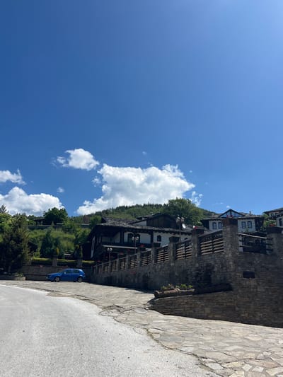The image captures a scene in Leshten, Bulgaria, featuring a narrow paved road running alongside stone architecture, indicative of traditional Bulgarian construction. A bright blue compact car is parked on the road's edge, close to a stone wall. The sky is a vibrant blue with scattered white clouds. The buildings in the background blend with the forested hillside, suggesting a village nestled in a mountainous region.