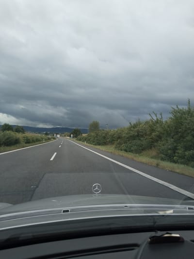 The image shows a view from inside a vehicle driving on a highway. The sky is heavily overcast with dark, stormy clouds. The road ahead curves to the left, with white lines marking the lanes. On either side of the road are dense green bushes and trees. In the distance, there are rolling hills or mountains. The hood of the car is visible in the foreground, with a Mercedes-Benz logo on it. The scene appears to be a highway in or near the city of Husa, Czechia, given the general landscape and infrastructure. There are no people or other vehicles clearly visible in the immediate foreground or on the road ahead. The overall impression is of a journey during inclement weather.