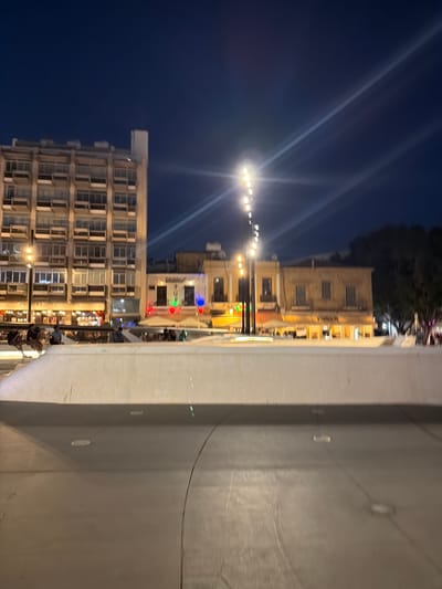 The image depicts an outdoor scene at night, possibly in Nicosia, Cyprus, given the context. The foreground is dominated by a concrete structure, possibly a wall or a raised platform, with visible circular indentations. To the left of this structure, a street or walkway is visible, lined with buildings.

Several buildings are present, appearing to be multi-story structures with numerous windows. Some buildings are illuminated from within, suggesting businesses or residences. String lights are strung across the area, adding to the nighttime ambiance. In the background, trees can be seen, their dark silhouettes against the night sky.

There are hints of activity. A few people are visible, some sitting at tables, possibly at outdoor dining establishments. The overall impression is that of an urban environment at night, with signs of public life and commerce. The time of day is clearly night, indicated by the dark sky and artificial lighting. No specific weather conditions are evident.

One building features signage with Greek text: "ΠΛΑΤΕΙΑ ΨΥΧΡΗ ΜΑΚΗΣ". This translates to "Psichri Maki Square". The presence of this sign strongly suggests the location is indeed a public square in Cyprus. The mood conveyed is one of urban evening leisure.