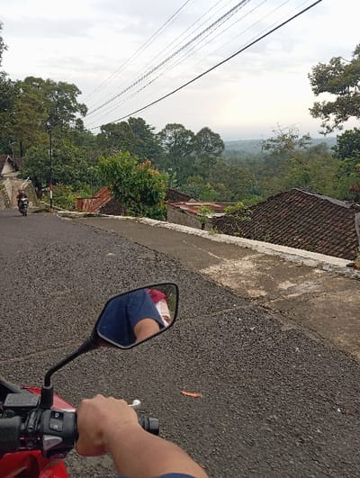 This image presents a scene from the perspective of a person operating a red motorcycle on an asphalt road in Bulukandang, Indonesia. In the immediate foreground, a human right hand, partially obscured by a dark blue sleeve, firmly grips the black handlebar grip. To the left, the motorcycle's black rearview mirror reflects a torso wearing a blue shirt, with a red and white patterned garment visible above. The dark gray asphalt road surface is coarse and exhibits minor cracks, with a single orange leaf resting on it.
The road ahead slopes downwards and curves gently to the left. A low concrete barrier or retaining wall runs along the right edge of the road, separating it from a downhill slope. Beyond this barrier, several structures with reddish-brown tiled roofs and at least one rusty corrugated metal roof are visible, partially integrated into the dense green foliage of the surrounding environment. A utility pole stands on the left side of the road, from which multiple black electrical wires span across the upper portion of the sky. Further along the road on the left, another motorcycle is observed, carrying two individuals; the passenger wears a visible red garment on their back. The background is dominated by an expansive view of lush green trees and dense vegetation covering hilly terrain, receding into a hazy distance under an overcast sky.