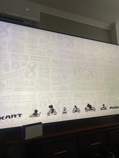 The image depicts a large display featuring a repeating "Mario Kart 8" themed wallpaper. The wallpaper includes various logos and imagery related to the video game. The display is positioned in a room, with a white air conditioner visible on the left. A Nintendo Switch console is placed at the bottom right corner of the display. A dark wooden dresser is visible to the right of the display. The exact location within La Aurora, Ecuador cannot be determined from this image.